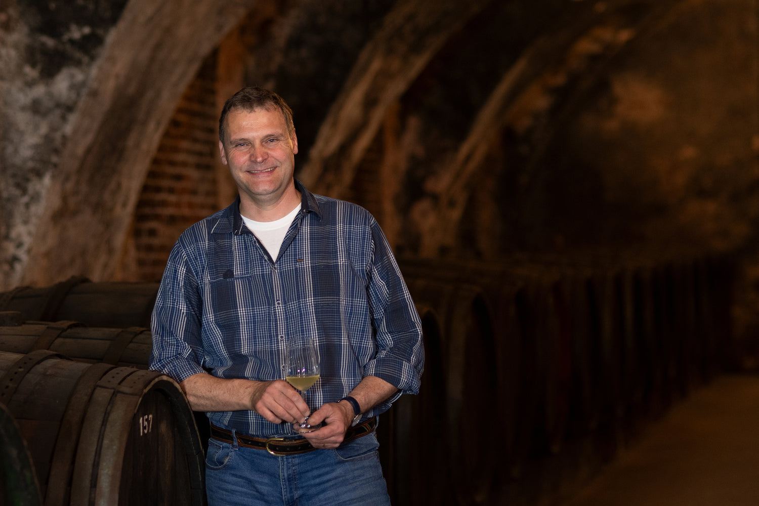 Mann mit kariertem Hemd steht lächelnd mit Weinglas im Weinkeller der Vereinigten Hospitien